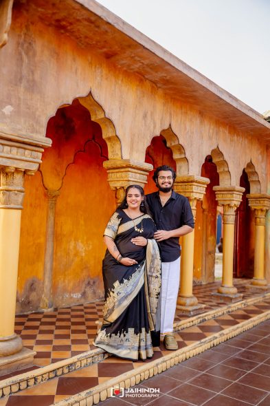 Pregnant woman in a black and gold saree poses with her partner under orange arches of a historic courtyard.