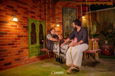 Couple relaxing on a wooden porch swing against a brick wall, smiling and looking at each other in warm evening light.