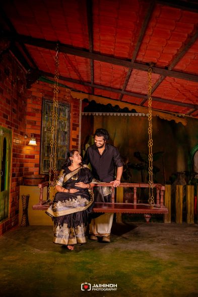 Couple sitting on a wooden porch swing indoors, smiling at each other in a brick-walled rustic setting.