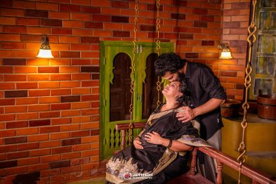 A couple shares a tender moment on a hanging bench in a warm brick-walled room, the man kissing the woman's forehead.