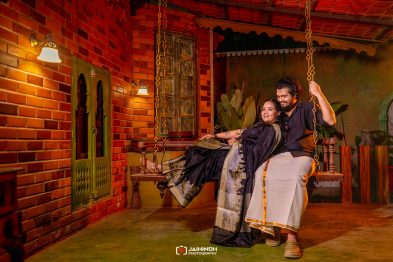 Couple in traditional attire smiling on a wooden swing inside a warmly lit brick courtyard.