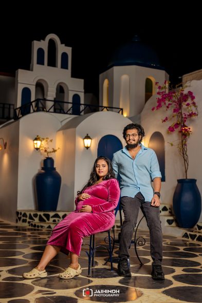 Pregnant woman in a pink velvet dress sits on a chair while a man stands beside her in a white-and-blue night-time courtyard with arches and blue domes.