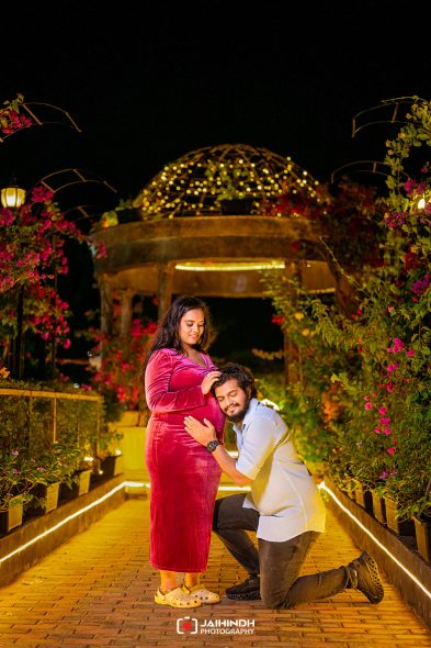 Pregnant woman in a red velvet dress stands as her partner kneels beside her, both in a flower-filled garden at night with warm lights.