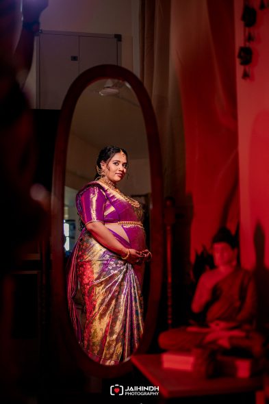 Pregnant woman in a shiny purple sari and gold jewelry stands near a mirror in a dim, red-lit room.
