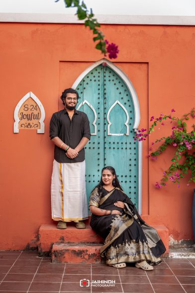 Couple in traditional attire posing in front of a turquoise arched door on an orange wall, with bougainvillea nearby.