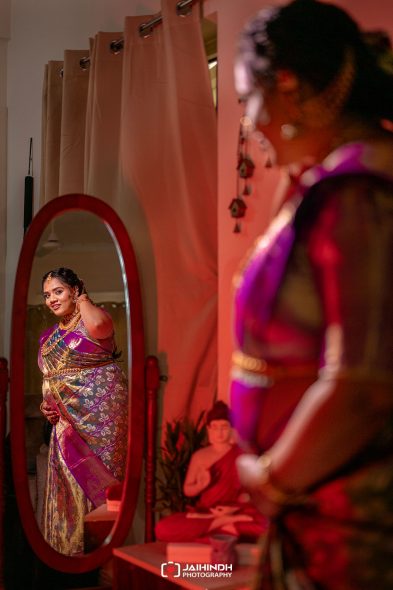 Woman in a purple-pink sari admires herself in a full-length mirror in a softly lit room with pink hues.
