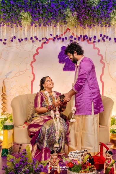 Bride seated on a beige sofa, wearing a purple-silver traditional saree and flower garlands, while a groom in a purple kurta offers a ring on a decorated stage with purple flowers overhead.