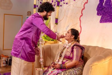 A groom in a purple kurta leans to kiss a smiling bride seated on a beige sofa, both dressed in purple wedding attire with floral garlands and jewelry in a decorated room.