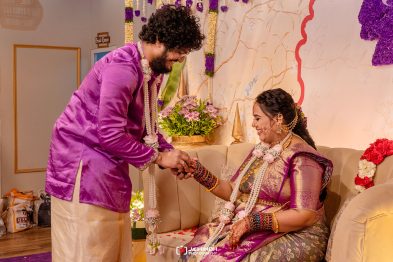 Groom in a magenta kurta helps place a ring on the bride’s finger as she sits smiling on a beige couch, surrounded by flowers and garlands at a wedding.