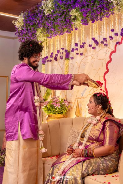 Groom in a purple kurta blesses the bride in a purple sari as she sits on a sofa, garlanded and smiling, during a wedding ceremony with purple and white flowers above.