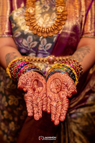 Bride's hands with intricate henna designs (mehndi), wearing colorful bangles and gold jewelry in a traditional wedding setting.