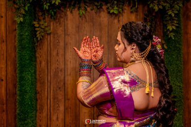 Bride in a purple saree with hennaed hands raised in prayer, ornate jewelry, and floral hair accessories against a wooden backdrop.