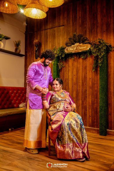 A man in a purple kurta gently holds a pregnant-looking woman in a colorful saree seated beside him, both smiling in a warm, festive setting with wood-paneled walls and decorative greenery behind them.
