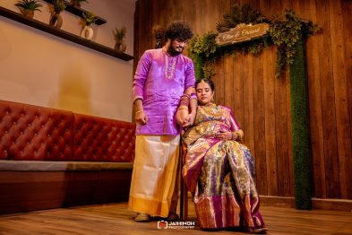 Man in a purple kurta stands beside a seated woman in a gold-purple saree, posing together at a wooden-walled wedding venue with greenery behind them.