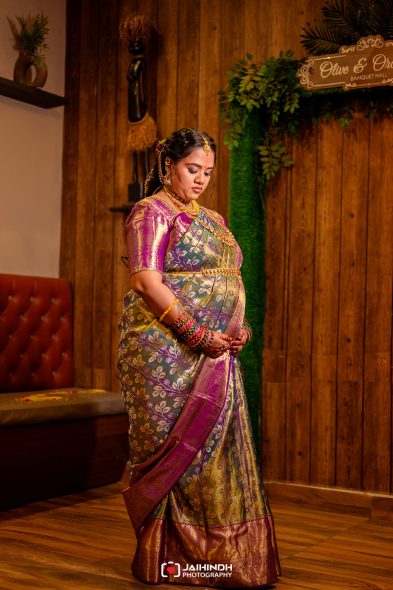 Pregnant woman in a colorful silk saree and heavy jewelry stands indoors, cradling her baby bump at a decorated event space.