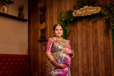 Pregnant woman in a colorful silk saree and heavy gold jewelry posing at a decorated banquet hall with a wooden wall backdrop and an 'Olive & Orchard' sign above.