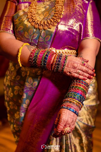 Close-up of arms crossed, wearing many colorful bangles and henna on hands against a purple and gold sari backdrop.