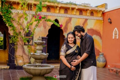 Expecting couple in a colorful courtyard; the man gently rests a hand on the woman’s baby bump as they smile together.