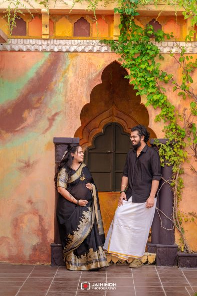 Pregnant woman in a black sari with gold detailing and her partner stand beside a colorful doorway with greenery, posing for a photo.