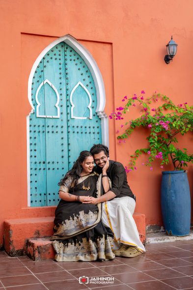 Couple sitting closely on orange steps beside a turquoise arched door, with pink flowers in a blue planter nearby.