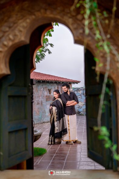Couple in traditional Indian attire stands back-to-back in a tiled courtyard, the woman resting her hand on her belly, framed by an ornate arched doorway.