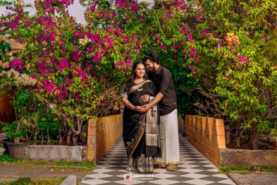 Pregnant woman in a black and gold saree stands with her partner on a checkered path, surrounded by bright pink bougainvillea flowers.