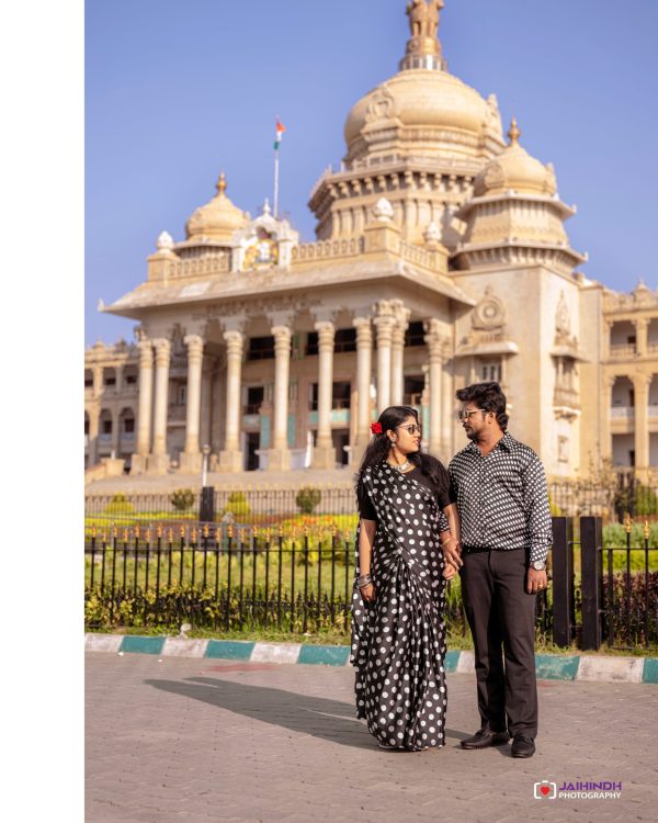 Couple wearing black-and-white polka dot outfits stands hand in hand in front of a grand beige temple with domes and a blue sky behind them.