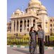 Couple wearing black-and-white polka dot outfits stands hand in hand in front of a grand beige temple with domes and a blue sky behind them.
