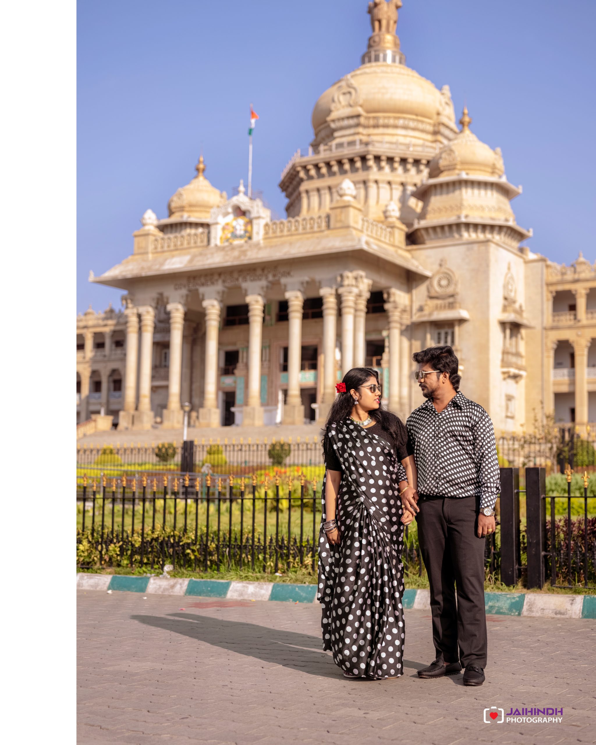Couple wearing black-and-white polka dot outfits stands hand in hand in front of a grand beige temple with domes and a blue sky behind them.