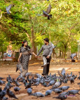Couple in black-and-white polka dot outfits holding hands while pigeons gather on a dirt path in a park with green trees in the background.