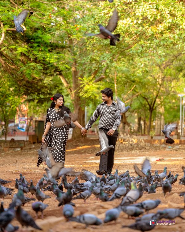 Couple in black-and-white polka dot outfits holding hands while pigeons gather on a dirt path in a park with green trees in the background.