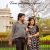 Couple in black-and-white polka dot outfits posing outdoors in a park, with a historic building and bare trees in the background.