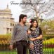 Couple in black-and-white polka dot outfits posing outdoors in a park, with a historic building and bare trees in the background.