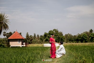 Bride in a pink sari stands in a green field as the groom, dressed in white, kneels and holds her hand with a small hut and palm trees in the background.