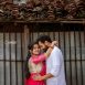 Couple embracing in front of a weathered tiled roof; woman in pink traditional attire and man in a white shirt.