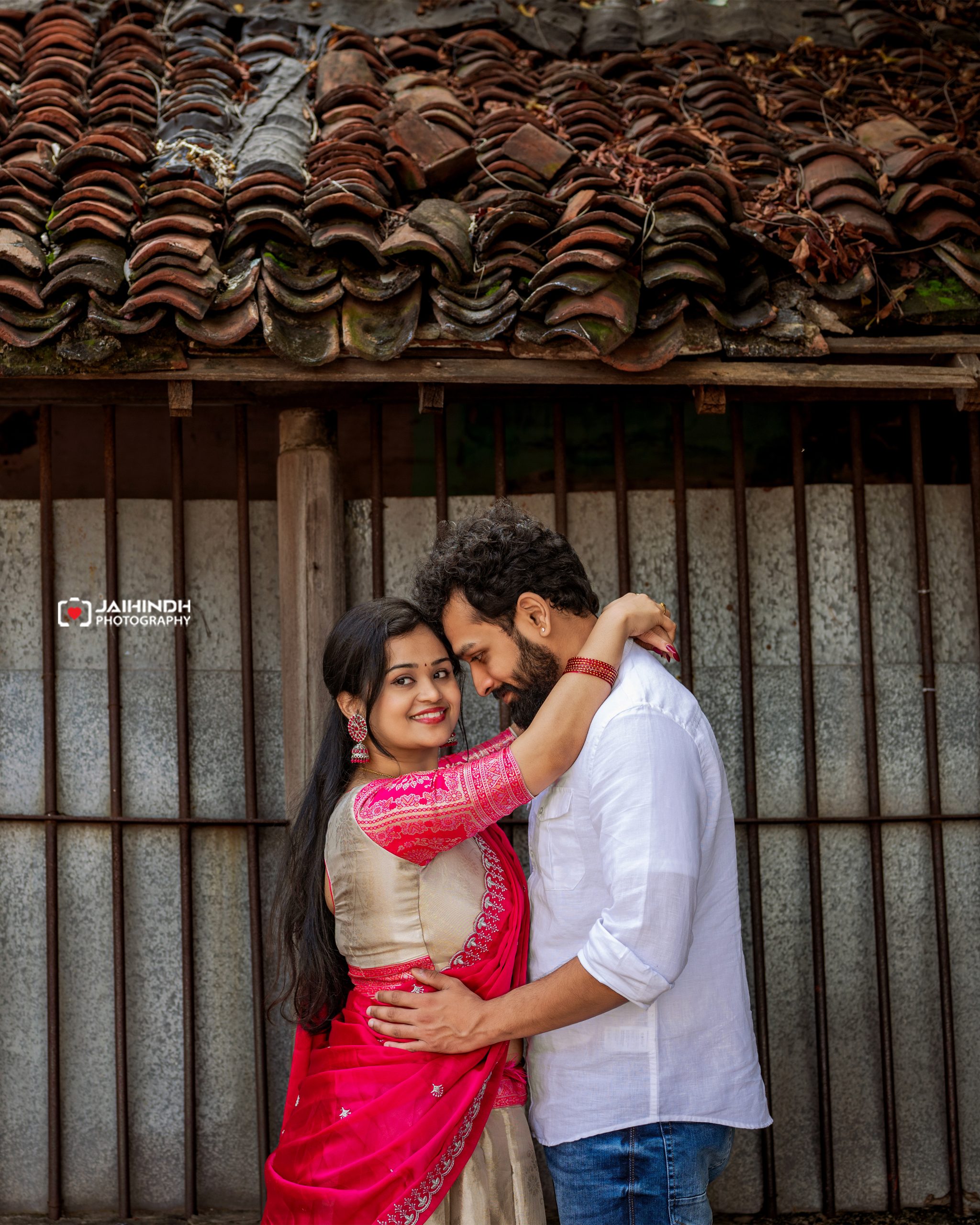 Couple embracing in front of a weathered tiled roof; woman in pink traditional attire and man in a white shirt.