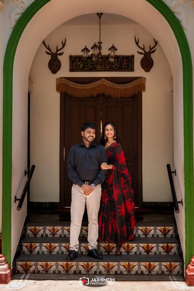 Couple posing on decorative tiled steps under a green arched doorway; the man wears a dark shirt and light pants while the woman wears a red and black saree, both smilingly facing the camera.