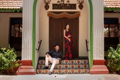 Couple posing on decorative tiled steps in front of a wooden double door; woman wears a red and black sari, man in a black shirt and light pants.