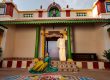 A man in a cream traditional outfit leans on a green column while a woman in a turquoise and yellow saree sits on the steps of a colorful temple entryway adorned with decorative tiles and arches.