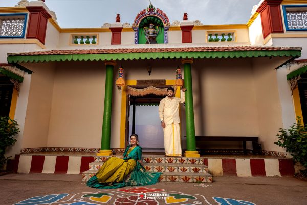 A man in a cream traditional outfit leans on a green column while a woman in a turquoise and yellow saree sits on the steps of a colorful temple entryway adorned with decorative tiles and arches.