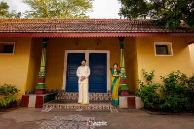 Couple in traditional attire posing on a colorful porch in front of a yellow house with blue doors and decorative green columns.