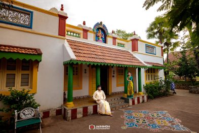 Colorful Indian house with decorative arches; a man in cream traditional attire sits on the porch while a woman in a green sari stands by a pillar; a rangoli design is on the ground.