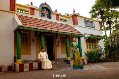 A man sits on the steps of a colorful temple-like building while a woman in a green and yellow sari stands nearby, both in traditional attire.