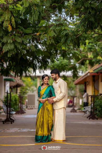 Romantic couple in traditional Indian attire stands close under a leafy tree in a courtyard, smiling at the camera side by side.