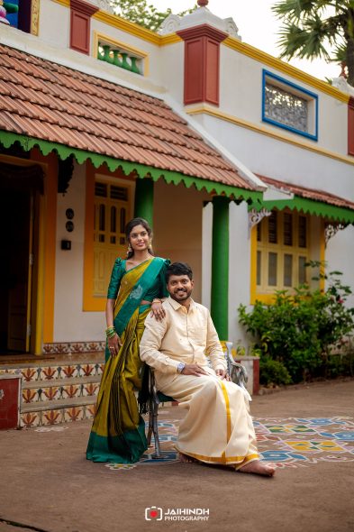 Couple in traditional Indian attire posing outside a colorful house with a tiled roof and patterned steps.