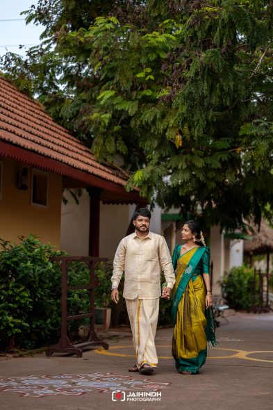 A couple walking hand in hand along a path outside a yellow building with green trees; man in cream kurta, woman in green-yellow saree wearing jewelry.