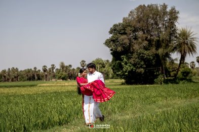 Couple in a green field; man lifts woman in a bright red saree as they smile at each other in an open rural setting.