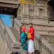 A couple in traditional Indian attire stands on a stone staircase with ornate temple architecture in the background, facing the camera warmly.