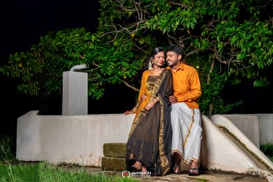 Couple in traditional Indian attire sit on a white wall at night, smiling at each other with green trees in the background.