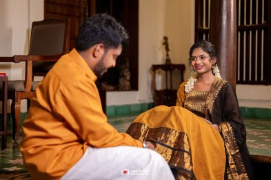 Woman in a gold and black sari sits on the floor, smiling at a man in an orange kurta indoors, with a jasmine garland in her hair.